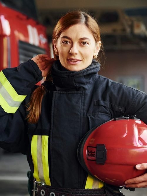 Holds red hat in hands. Female firefighter in protective uniform standing near truck