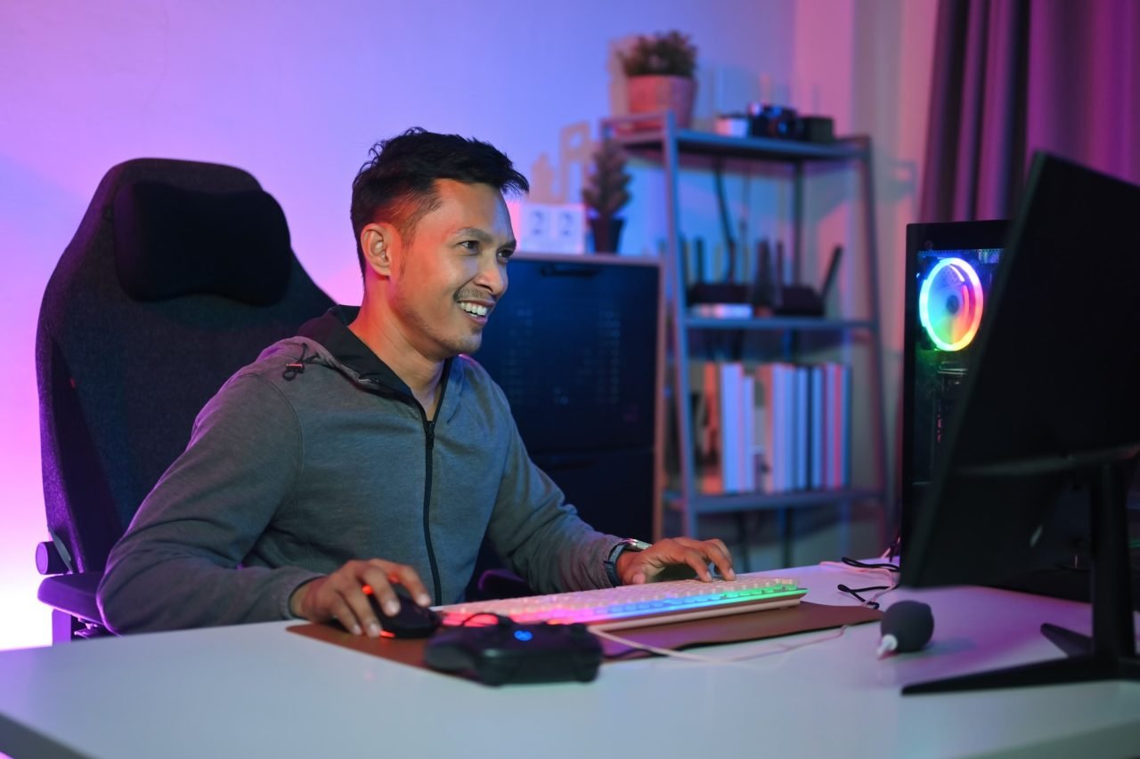 Smiling Asian male gamer playing video game at gaming room at night.