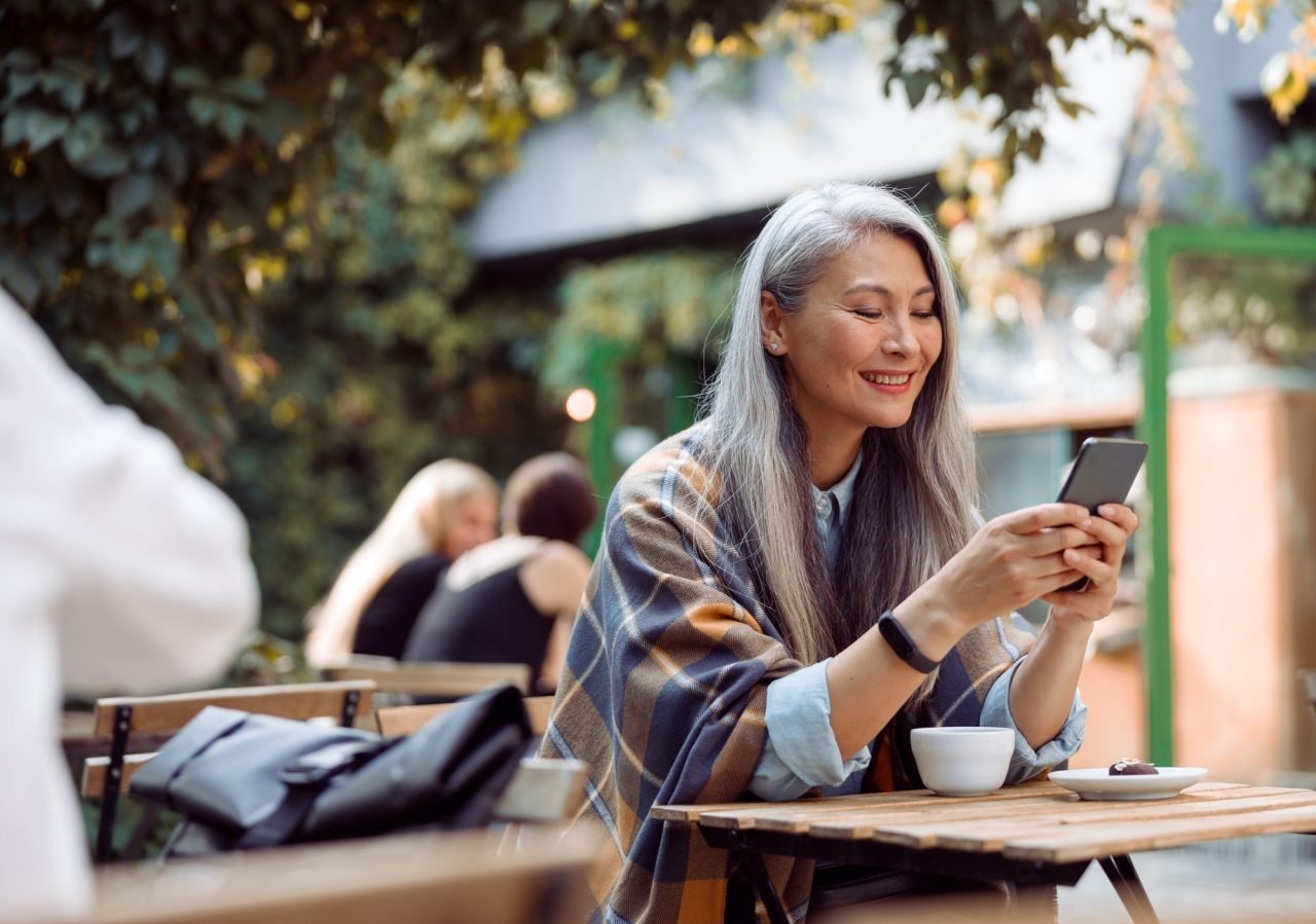Happy mature Asian woman surfs internet on phone at small table on outdoors cafe terrace