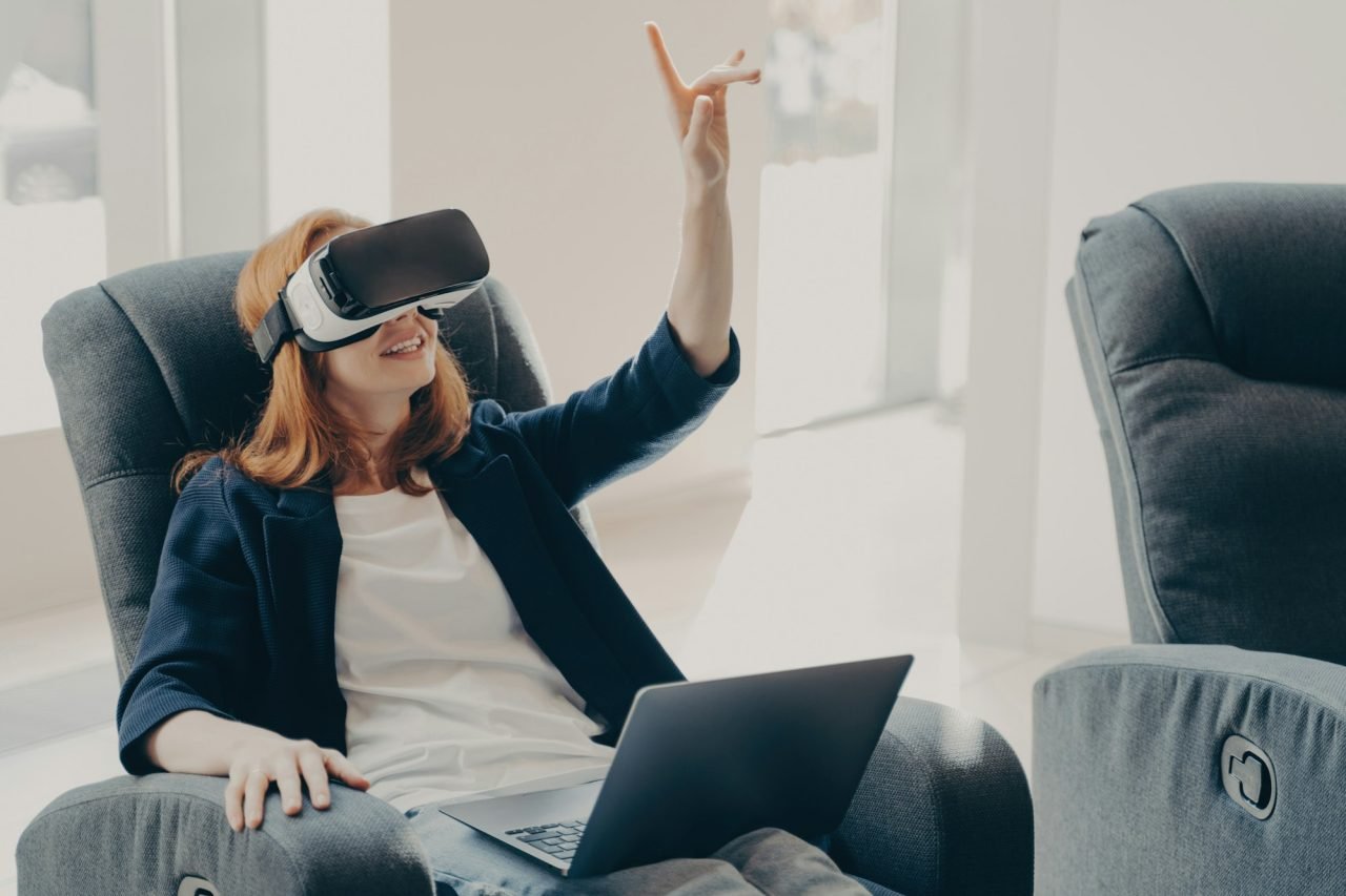 Female in VR headset pointing with finger, touching 3d object while working remotely on laptop