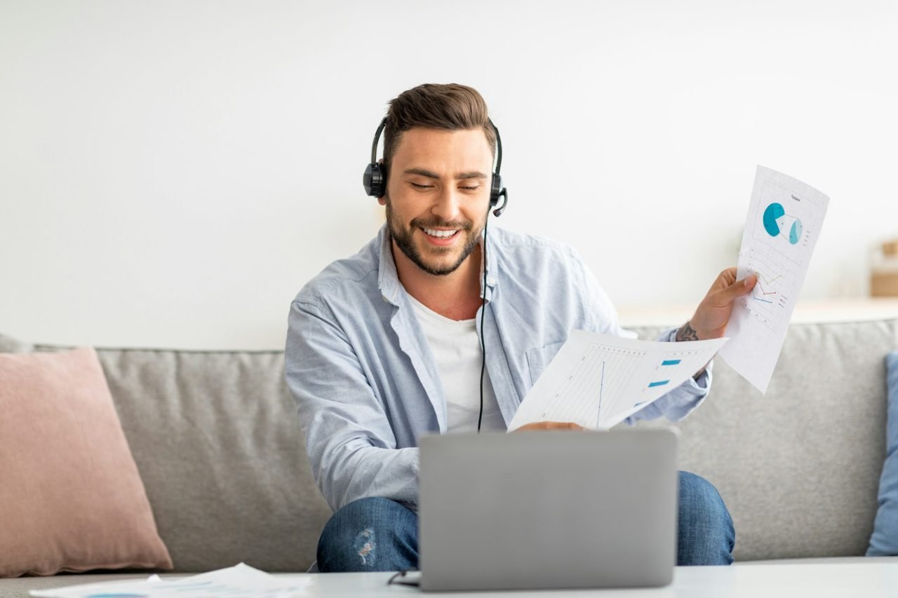 Virtual meeting. Happy man having video call, showing documents with diagrams to laptop camera